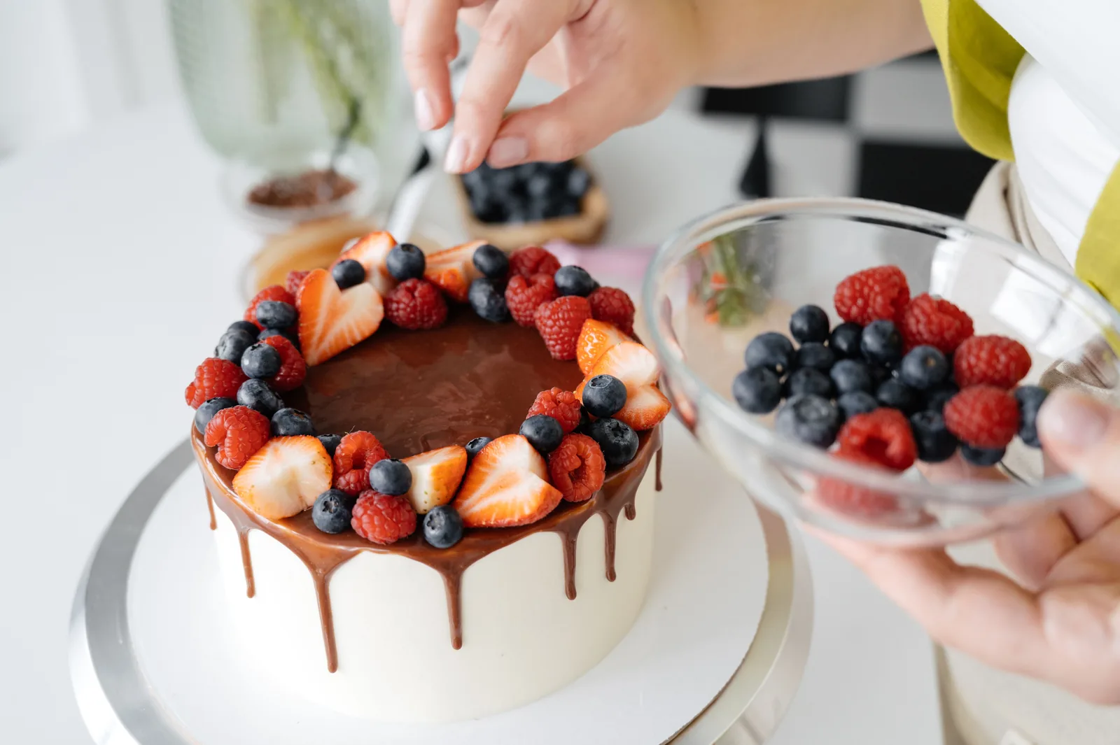 Hands placing strawberries and blueberries on a frosted cake